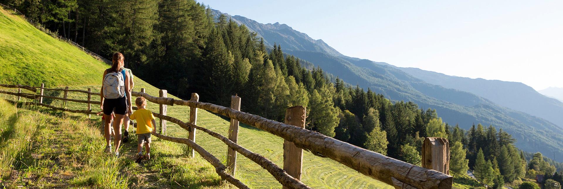 Eine Mutter wandert mit ihrem Kind auf Almweg mit Holzzaun und Blick auf bewaldete Berge im Abendlicht.